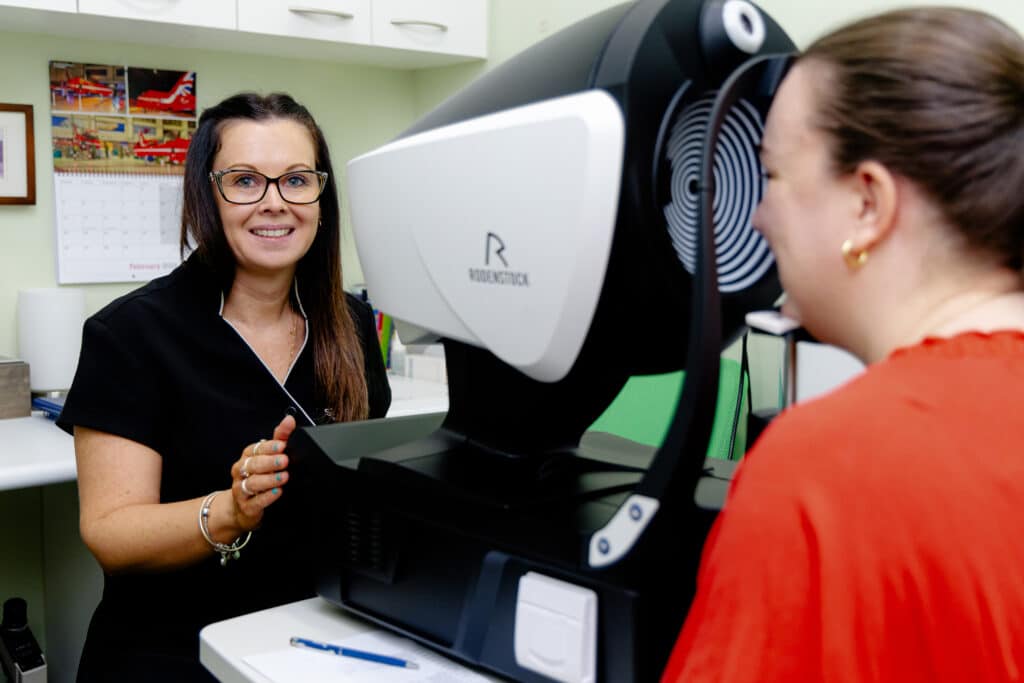 A woman looking into an eye testing machine while another woman looks onward smiling.